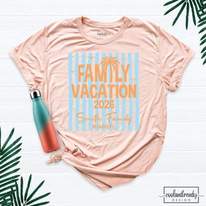 May include: A peach-colored t-shirt featuring a graphic with light blue vertical stripes, an orange palm tree, and the text "FAMILY VACATION 2026 Smith Family MIAMI FL". A teal and orange gradient water bottle is positioned on the shirt.