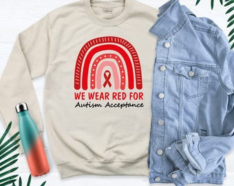 May include: A beige crewneck sweatshirt featuring a red and pink rainbow graphic with a red awareness ribbon and the text WE WEAR RED FOR Autism Acceptance. A light blue denim jacket and a teal and orange gradient water bottle are nearby.