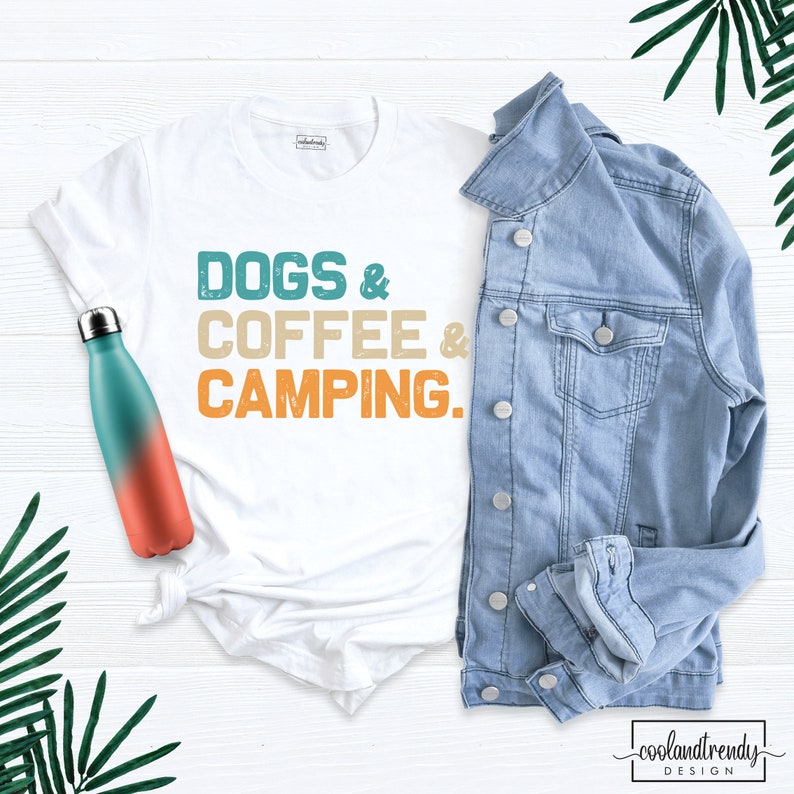 Pu&ograve; includere: Maglietta bianca con il testo "DOGS & COFFEE & CAMPING." in un font di stile vintage. Il testo &egrave; in tonalit&agrave; di turchese, arancione e beige. La maglietta &egrave; appoggiata su una superficie di legno bianco con una giacca di jeans blu e una bottiglia d'acqua turchese e arancione.