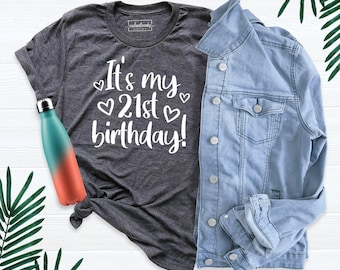 May include: A dark gray short-sleeved t-shirt with white text that reads It's my 21st birthday! surrounded by small heart outlines, paired with a light blue denim jacket and a teal and orange gradient metal water bottle.