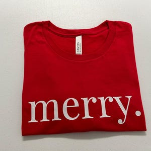Puede incluir: Una camiseta roja doblada con la palabra "merry." impresa en blanco. La camiseta est&aacute; hecha de un material suave y tiene cuello redondo. La etiqueta dice "Canvas".