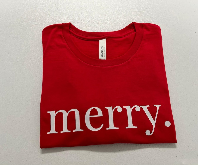Puede incluir: Una camiseta roja doblada con la palabra "merry." impresa en blanco. La camiseta est&aacute; hecha de un material suave y tiene cuello redondo. La etiqueta dice "Canvas".