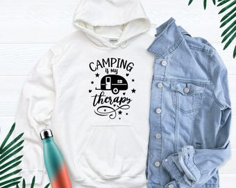 May include: A white hooded sweatshirt featuring a black graphic of a camper trailer with the text Camping is my therapy, accompanied by a light blue denim jacket and a teal and orange gradient metal water bottle.