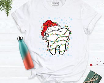 May include: A white short-sleeve t-shirt featuring a graphic of a tooth wearing a red Santa hat and wrapped in colorful Christmas string lights, surrounded by light blue snowflakes. The shirt has a small tag at the neck labeled Cookstrendy Design.