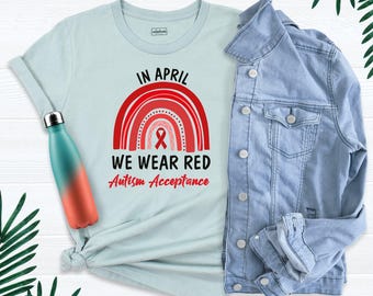 May include: A light blue t-shirt featuring a red rainbow graphic with a red awareness ribbon and the text "IN APRIL WE WEAR RED Autism Acceptance" in black and red, accompanied by a teal and orange gradient water bottle and a light blue denim jacket.