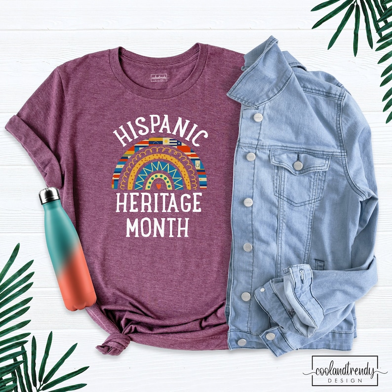 May include: A maroon t-shirt with a white graphic design that says "Hispanic Heritage Month". The graphic features a rainbow-colored arch with flags of different Hispanic countries.