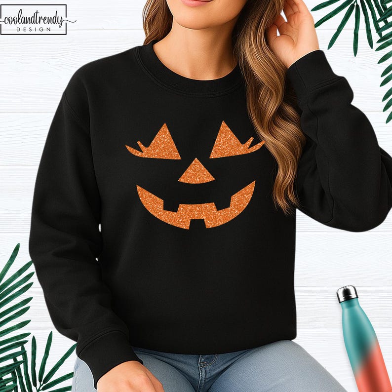 May include: Black sweatshirt featuring a glittery orange jack-o'-lantern face design. The design includes triangular eyes, a triangular nose, and a wide, smiling mouth. The sweatshirt has a crew neck and long sleeves. The brand name "coolandtrendy" is in the top left corner.