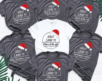 May include: A collection of six t-shirts, five dark heather gray and one white, each featuring a circular graphic with a red and white Santa hat. The text reads: Most Likely To Shake the presents, Shop for Christmas, Drink all the wine, Return all the gifts, Sing Christmas songs, Eat Santa's cookies, and Stay home.