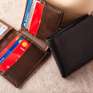 May include: A brown leather bi-fold wallet is open, displaying credit cards and an ID. A closed black leather wallet sits next to it. The wallets are on a neutral surface. The open wallet shows multiple credit cards.