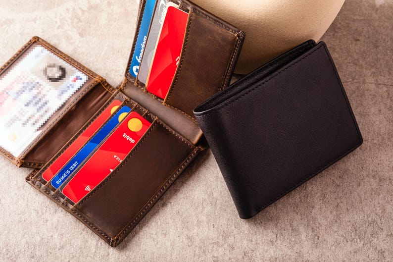 May include: A brown leather bi-fold wallet is open, displaying credit cards and an ID. A closed black leather wallet sits next to it. The wallets are on a neutral surface. The open wallet shows multiple credit cards.