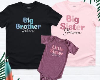 May include: A black t-shirt with blue text reading Big Brother Robert, a light pink t-shirt with pink text reading Big Sister Sharon, and a maroon baby bodysuit with pink text reading Little Sister Almira, plus a gradient water bottle.