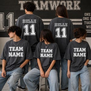 K&ouml;nnte beinhalten: Vier anthrazitfarbene T-Shirts mit wei&szlig;er Blockschrift. Zwei T-Shirts zeigen den Text "BELLE 11" auf der R&uuml;ckseite, w&auml;hrend alle vier T-Shirts den Text "TEAM NAME" auf der Vorderseite tragen. Die T-Shirts werden mit hellblauen Jeans kombiniert.