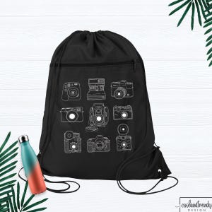 May include: A black drawstring backpack featuring a white line art graphic of nine different vintage cameras arranged in a grid. The bag has a zippered front pocket and black cord straps. A teal and orange gradient water bottle is nearby.