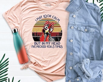 May include: A peach-colored t-shirt featuring a graphic of a chicken wearing a red polka-dot headband, with the text I MAY LOOK CALM BUT IN MY HEAD I'VE PECKED YOU 3 TIMES, alongside a teal and orange gradient water bottle and a light blue denim jacket.