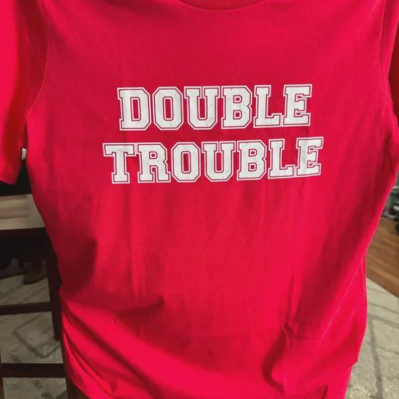 K&ouml;nnte beinhalten: Ein leuchtend rotes Kurzarm-T-Shirt mit dem Text DOUBLE TROUBLE, der in wei&szlig;en Blockbuchstaben auf der Brust aufgedruckt ist. Die Schrift hat ein leicht abgenutztes, verwittertes Aussehen und das T-Shirt hat einen klassischen Rundhalsausschnitt.