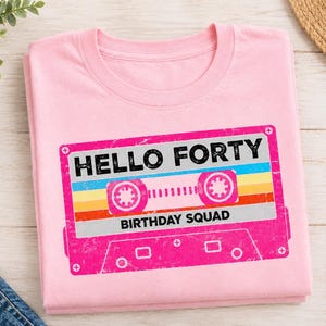 Pu&ograve; includere: Una t-shirt rosa chiaro con la grafica di una cassetta audio retr&ograve; nei toni del rosa, giallo e blu. Il testo "HELLO FORTY" &egrave; stampato in nero sopra la cassetta, e "BIRTHDAY SQUAD" &egrave; stampato in nero sotto la grafica della cassetta.