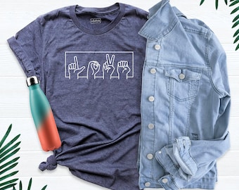May include: A heather navy blue t-shirt featuring a white line art graphic of four hands spelling the word LOVE in American Sign Language, paired with a light blue denim jacket and a teal and coral gradient metal water bottle.
