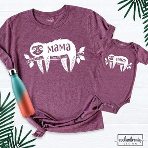 May include: Burgundy t-shirt and onesie set with a white sloth design on a branch. The t-shirt has the word "Mama" and the onesie has the word "Baby". A teal and orange water bottle is on the left side of the image.