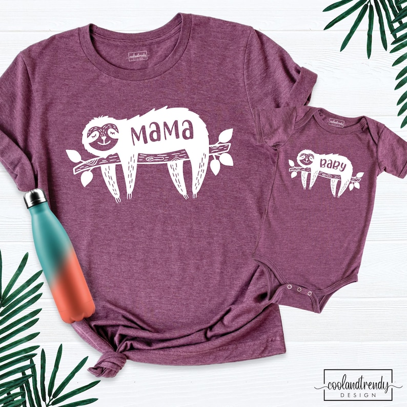 May include: Burgundy t-shirt and onesie set with a white sloth design on a branch. The t-shirt has the word "Mama" and the onesie has the word "Baby". A teal and orange water bottle is on the left side of the image.