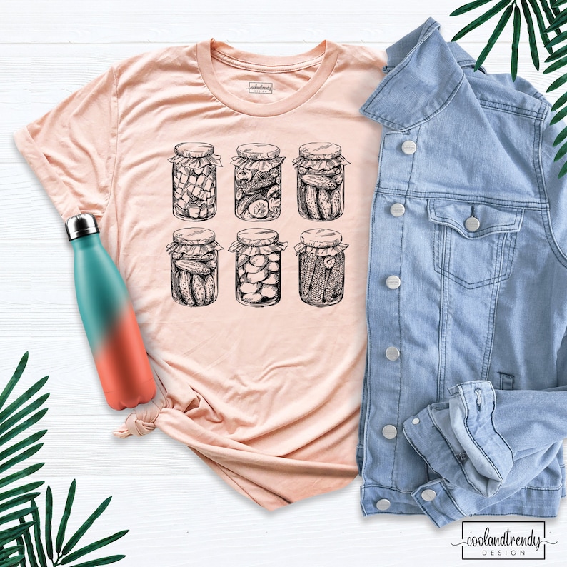 Puede incluir: Una camiseta color melocot&oacute;n con una ilustraci&oacute;n en blanco y negro de seis tarros de pepinillos. Una botella de agua turquesa y coral y una chaqueta vaquera azul claro tambi&eacute;n est&aacute;n en la imagen. El texto "coolandtrendy design" est&aacute; en la parte inferior.