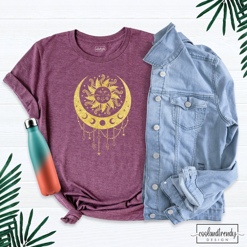 Puede incluir: Una camiseta color granate con un dise&ntilde;o de sol y luna en purpurina dorada, con forma de luna creciente y una cara en el sol. El dise&ntilde;o incluye una cadena colgante de estrellas y diamantes.