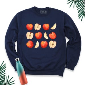 May include: A navy blue crewneck sweatshirt featuring a repeating pattern of red apples and apple slices across the chest. The interior neck label displays the text "coolandtrendy DESIGN". A gradient teal and orange water bottle is positioned below.