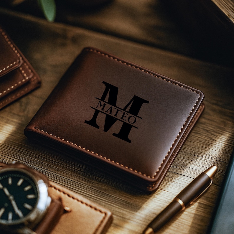 May include: A brown leather wallet with the name "MATEO" and the initial "M" engraved on the front. The wallet is rectangular and has a stitched border. A watch and pen are visible in the background.
