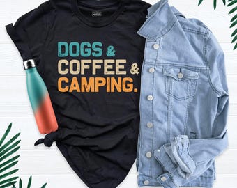 May include: A black short-sleeve t-shirt with the text DOGS & COFFEE & CAMPING in teal, beige, and orange. A teal and orange gradient water bottle is placed on the shirt, and a light blue denim jacket is layered next to the t-shirt.
