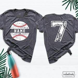 May include: A dark heather gray short-sleeve t-shirt featuring a white baseball graphic with red stitching and the word NAME on the front, and a large white number 7 with baseball stitching details on the back of the garment.