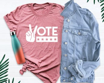 May include: A heathered mauve t-shirt featuring a white graphic of a hand making a peace sign next to the word VOTE and five stars. Beside the shirt is a light blue denim jacket and a teal and orange gradient metal water bottle.