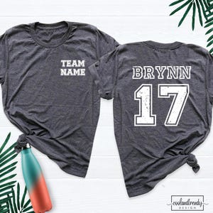 May include: A dark heather gray short-sleeve t-shirt featuring the text TEAM NAME on the front left chest and the name BRYNN with the number 17 in large white distressed lettering on the back. The shirt has a crew neckline.