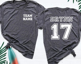May include: A dark heather gray short-sleeve t-shirt featuring the text TEAM NAME on the front left chest and the name BRYNN with the number 17 in large white distressed lettering on the back. The shirt has a crew neckline.