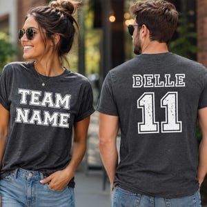 K&ouml;nnte beinhalten: Ein dunkel anthrazitfarbenes Kurzarm-T-Shirt. Die Vorderseite zeigt den wei&szlig;en Schriftzug "TEAM NAME" in einer College-Blockschrift. Die R&uuml;ckseite zeigt den wei&szlig;en Schriftzug "BELLE" &uuml;ber der gro&szlig;en Zahl "11" im gleichen College-Stil.