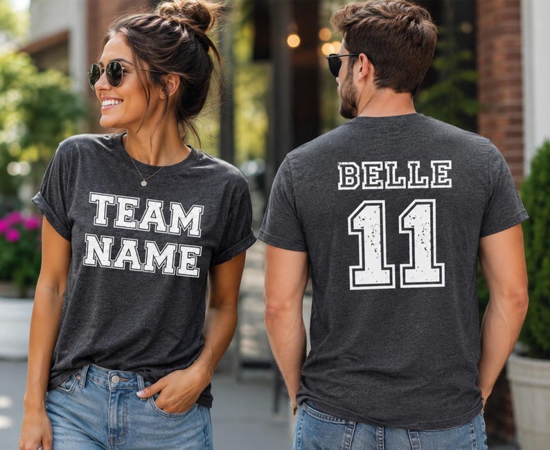 K&ouml;nnte beinhalten: Ein dunkel anthrazitfarbenes Kurzarm-T-Shirt. Die Vorderseite zeigt den wei&szlig;en Schriftzug "TEAM NAME" in einer College-Blockschrift. Die R&uuml;ckseite zeigt den wei&szlig;en Schriftzug "BELLE" &uuml;ber der gro&szlig;en Zahl "11" im gleichen College-Stil.