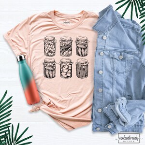 Puede incluir: Una camiseta de color melocot&oacute;n claro con una ilustraci&oacute;n en blanco y negro de seis tarros de pepinillos. Una botella de agua turquesa y coral y una chaqueta vaquera azul claro tambi&eacute;n est&aacute;n en la imagen. La camiseta tiene el texto "coolandtrendy design".