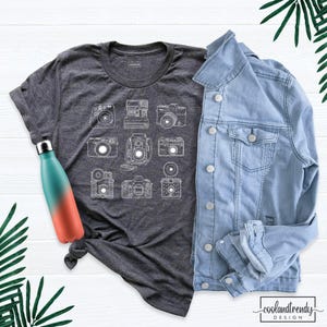 May include: A dark gray short-sleeve t-shirt featuring a white line art graphic of nine different vintage cameras. A light blue denim jacket is layered next to the shirt, and a teal and coral gradient metal water bottle is placed on the left.