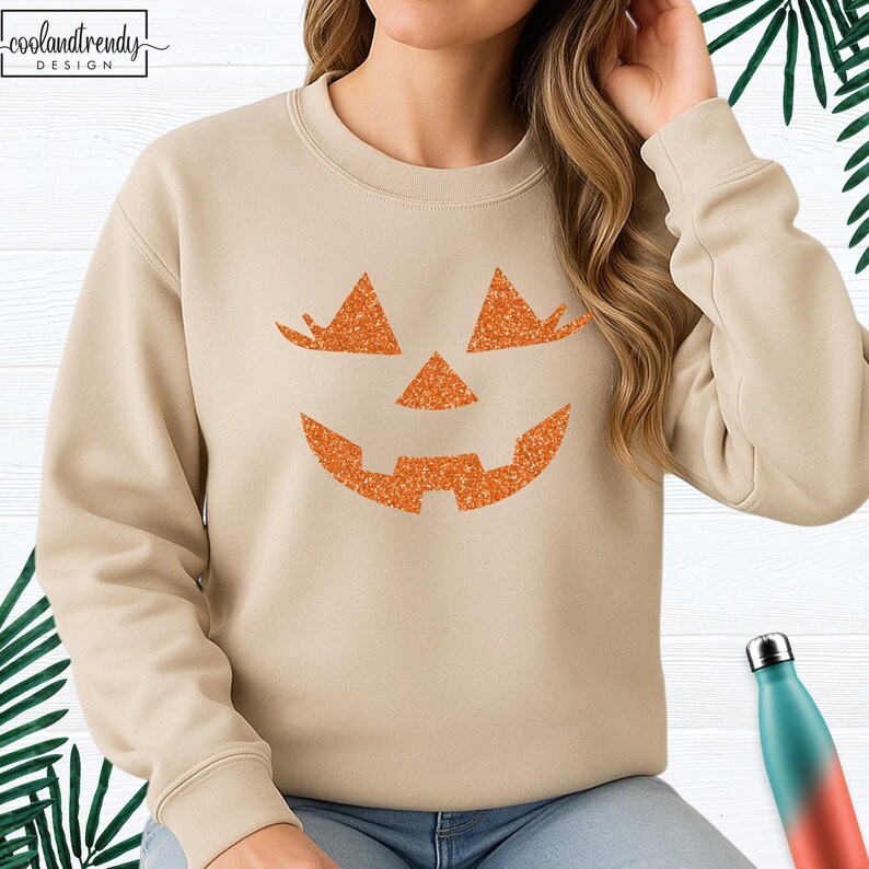 May include: Beige sweatshirt featuring a glittery orange jack-o'-lantern face design. The sweatshirt has a crew neck and long sleeves. The text "coolandtrendy DESIGN" is in the upper left corner. A teal and red water bottle is in the lower right corner.