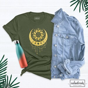 Puede incluir: Una camiseta verde con un dise&ntilde;o de sol y luna dorado. El dise&ntilde;o presenta un sol con cara y una luna creciente con estrellas y diamantes colgando de ella.