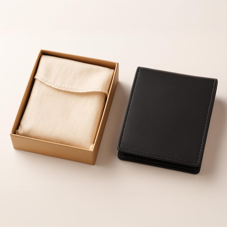 May include: A black leather wallet with a stitched edge is positioned beside a tan cardboard box holding a folded, cream-coloured cloth. The wallet is rectangular and closed. The box is open, showing the cloth.