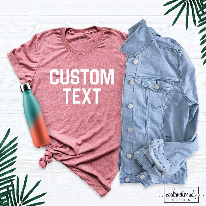 May include: A pink t-shirt with the text "CUSTOM TEXT" in white letters. The t-shirt is laid flat on a white wooden surface with a denim jacket and a water bottle.
