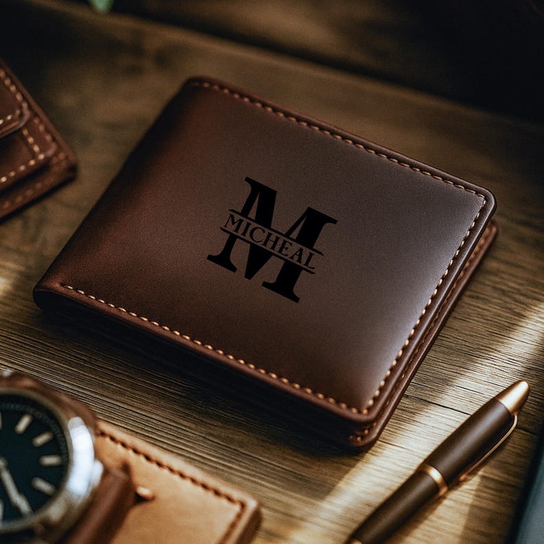 May include: A brown leather wallet with the name "Micheal" and the letter "M" engraved on the front. The wallet is rectangular and has a stitched border. A watch and pen are visible in the background.