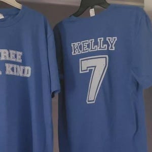 K&ouml;nnte beinhalten: Zwei blaue kurz&auml;rmelige T-Shirts, die an Kleiderb&uuml;geln h&auml;ngen. Das linke T-Shirt zeigt teilweise sichtbaren wei&szlig;en Text. Das rechte T-Shirt zeigt den wei&szlig;en Text KELLY &uuml;ber der gro&szlig;en wei&szlig;en Zahl 7, beide mit einer abgenutzten Textur.