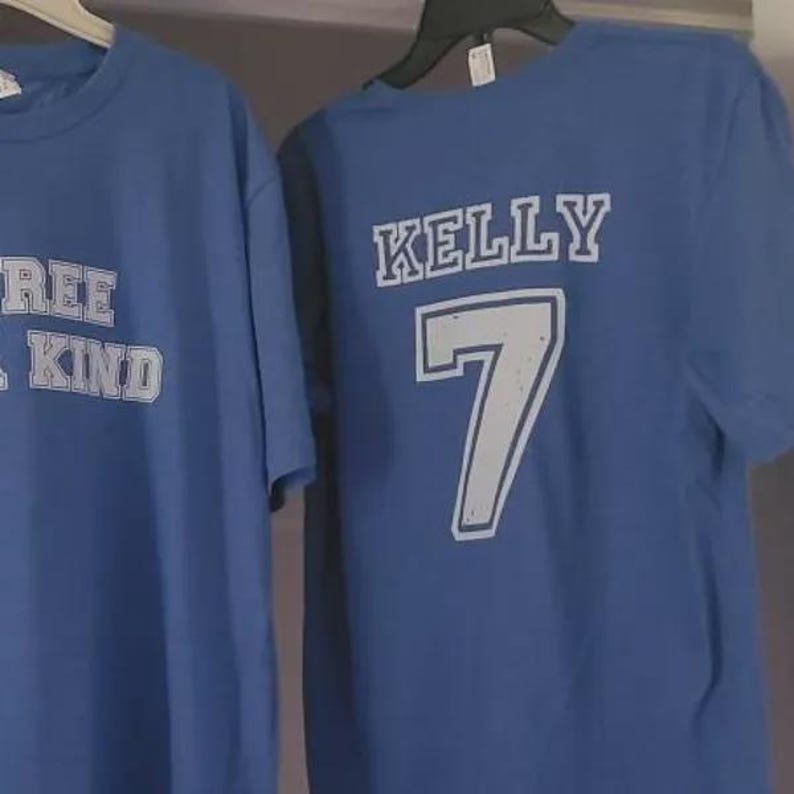 K&ouml;nnte beinhalten: Zwei blaue kurz&auml;rmelige T-Shirts, die an Kleiderb&uuml;geln h&auml;ngen. Das linke T-Shirt zeigt teilweise sichtbaren wei&szlig;en Text. Das rechte T-Shirt zeigt den wei&szlig;en Text KELLY &uuml;ber der gro&szlig;en wei&szlig;en Zahl 7, beide mit einer abgenutzten Textur.