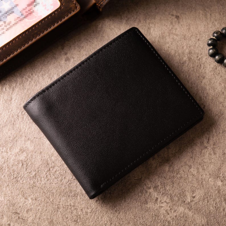May include: A black leather wallet with a smooth finish. The rectangular bi-fold wallet has visible stitching around the edges. A section of a brown leather wallet is in the background. The wallet is likely to be 10-12 cm wide.