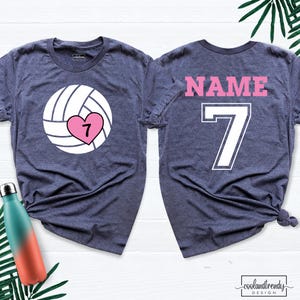 May include: Two navy blue t-shirts. The front shirt has a volleyball graphic with a pink heart and the number 7. The back shirt has the word "NAME" and the number 7 in pink and white. The shirts are from coolandtrendy design.