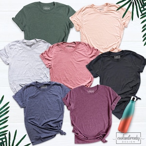 Op de afbeelding: Zeven blanco t-shirts in verschillende kleuren, waaronder groen, roze, grijs, rood, zwart, blauw en paars, gelegd op een wit houten oppervlak. De shirts zijn allemaal met ronde hals en korte mouwen.
