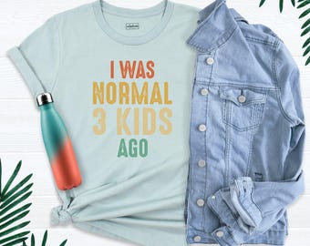 May include: A light blue t-shirt featuring the text I WAS NORMAL 3 KIDS AGO in orange, yellow, and green. The shirt is paired with a light blue denim jacket and a teal and orange gradient water bottle, all arranged on a white surface.