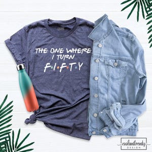 May include: A dark heather blue t-shirt with white text that reads THE ONE WHERE I TURN F I F T Y, featuring small colored dots between the letters. Next to it is a light blue denim jacket and a teal and coral gradient metal water bottle.
