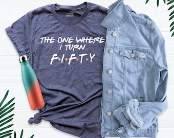 May include: A dark heather blue t-shirt with white text that reads THE ONE WHERE I TURN F I F T Y, featuring small colored dots between the letters. Next to it is a light blue denim jacket and a teal and coral gradient metal water bottle.