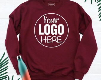 May include: A maroon long-sleeve crewneck sweatshirt featuring a white circular graphic with the text "YOUR LOGO HERE" in the center. A gradient teal and orange metal water bottle is positioned in the lower left corner of the frame.
